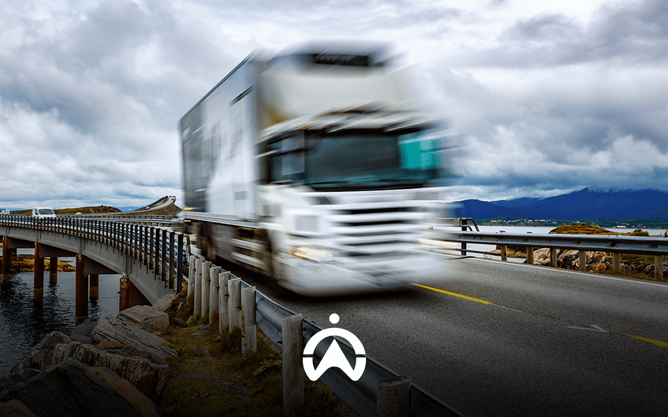 A blurred white truck speeding on a bridge over water, against a backdrop of dramatic clouds and distant mountains.
