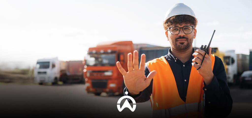 A construction worker in an orange vest and hard hat holds a walkie-talkie, signaling to stop with an outstretched hand, surrounded by trucks.