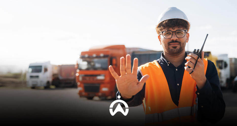 A construction worker in an orange vest and hard hat holds a walkie-talkie, signaling to stop with an outstretched hand, surrounded by trucks.