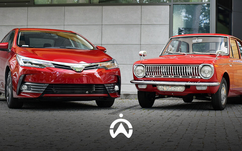 A modern red sedan and a vintage red car parked side by side against a neutral stone background, showcasing contrasting automotive designs.