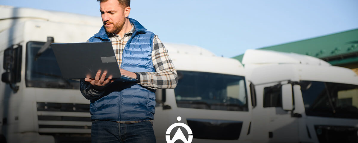 A person in a blue vest stands outdoors, working on a laptop, with large white trucks observed in the background.