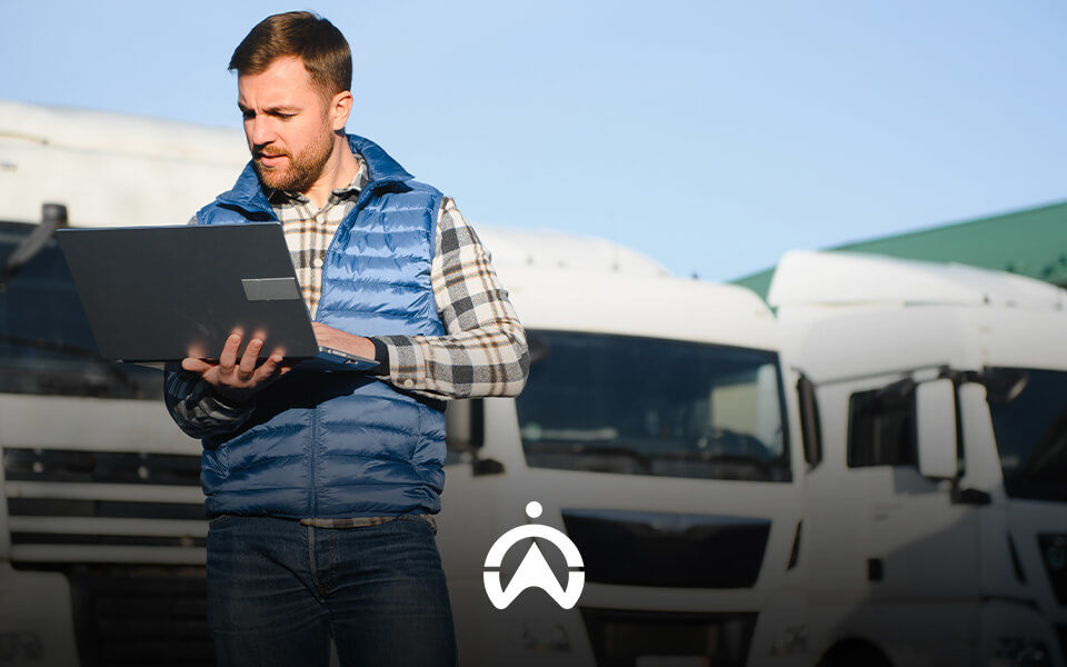 A person in a blue vest stands outdoors, working on a laptop, with large white trucks observed in the background.