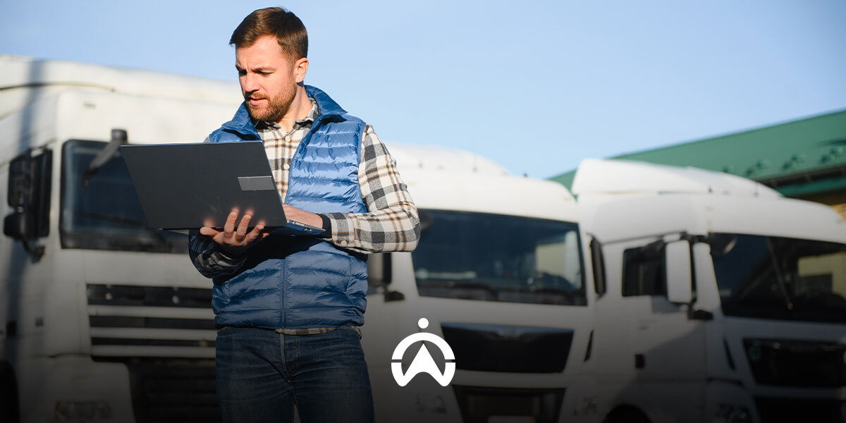 A person in a blue vest stands outdoors, working on a laptop, with large white trucks observed in the background.