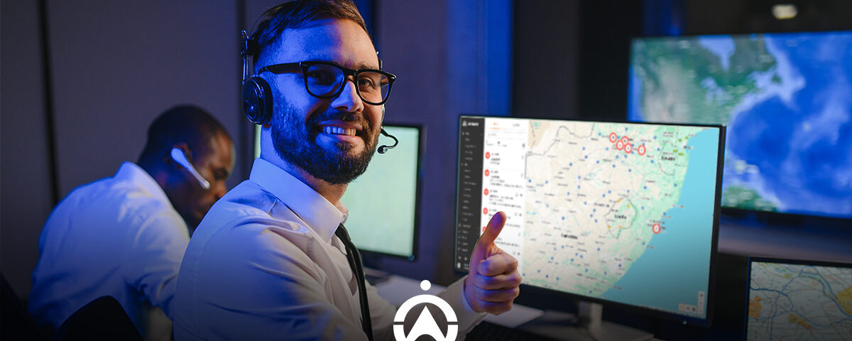 A man wearing a headset gives a thumbs-up in a high-tech control room filled with monitors displaying maps and data.