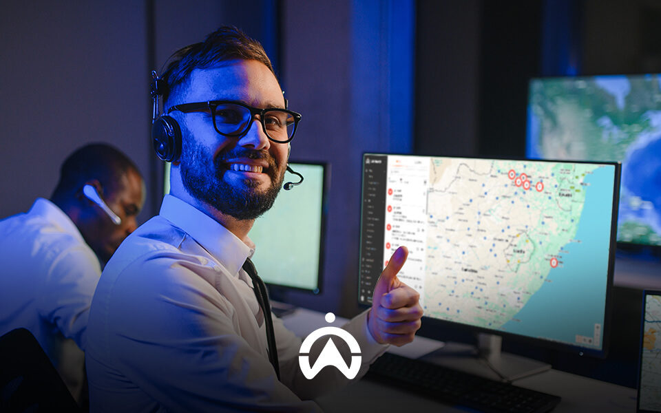 A man wearing a headset gives a thumbs-up in a high-tech control room filled with monitors displaying maps and data.